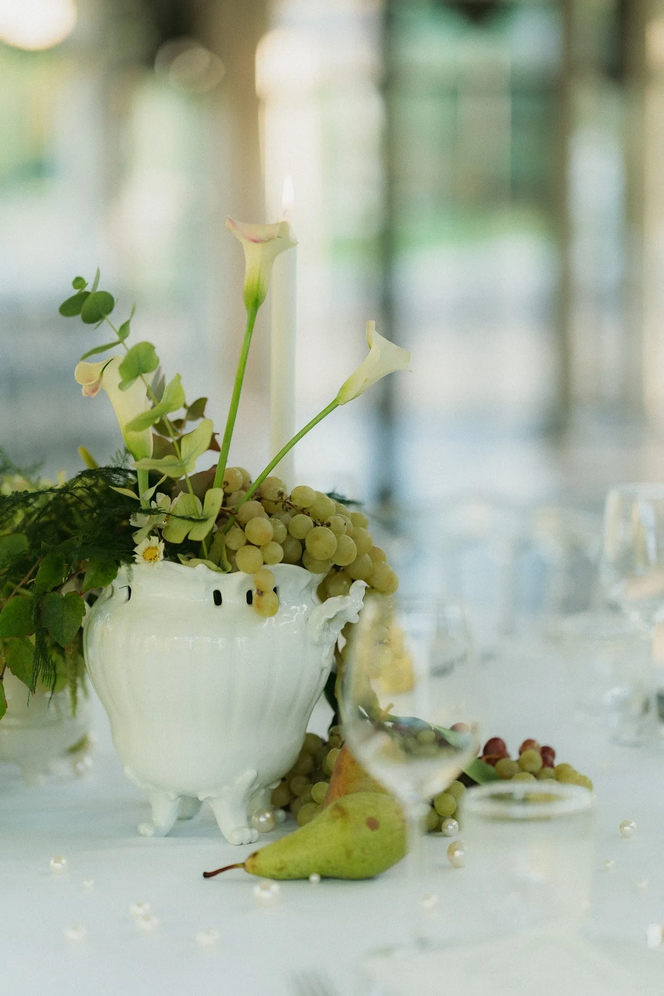 Vase blanc garni de raisins, d'arums et de feuillages, une poire posée à côté