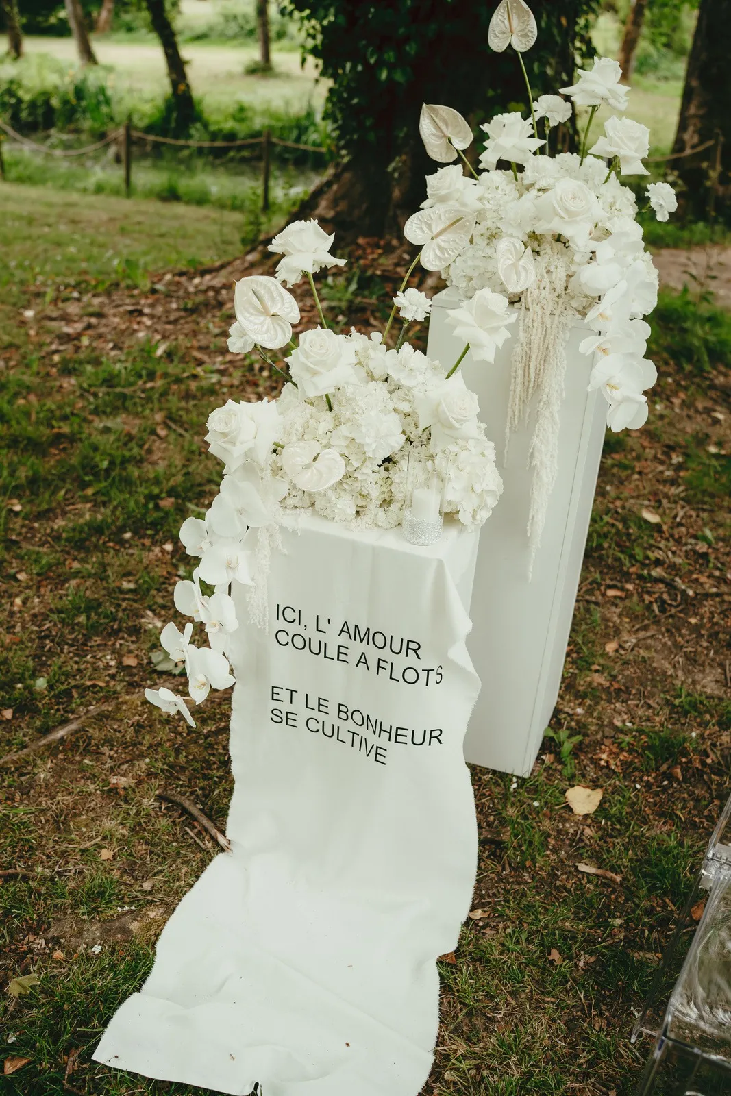 Colonnes blanches fleuries de roses et d'orchidées, avec un voile brodé « Ici, l'amour coule à flots »