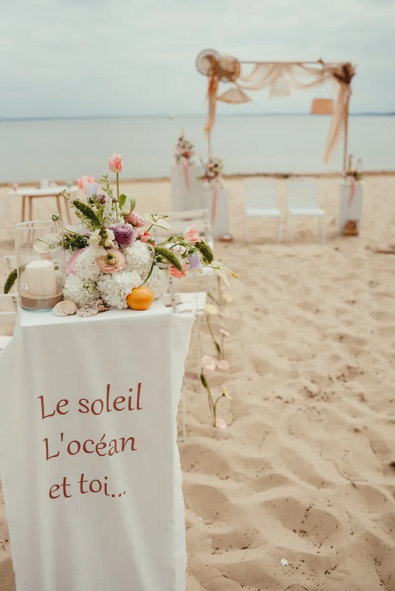Arche de cérémonie sur le sable, fleurs pastel et bannière « Le soleil, l'océan et toi »